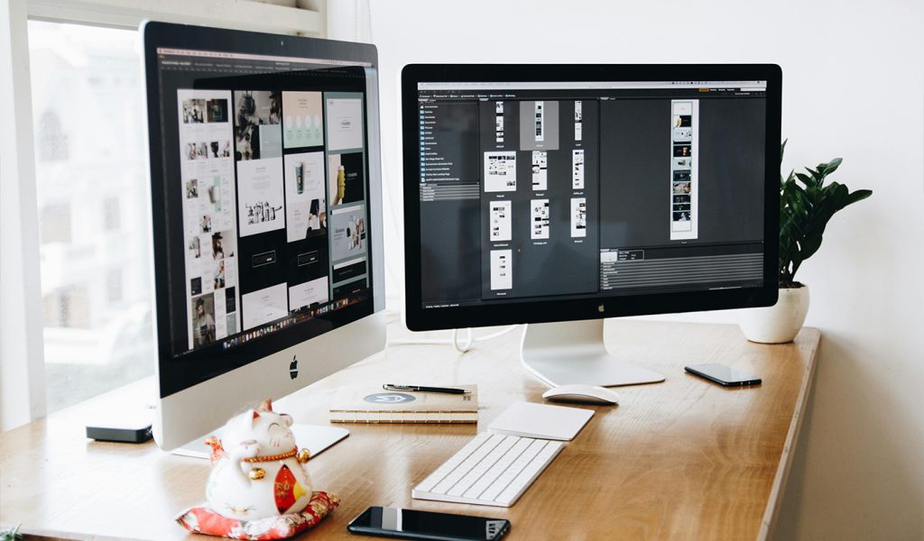 Two computer screens with design software open, with a person working to build an online presence for their book launch